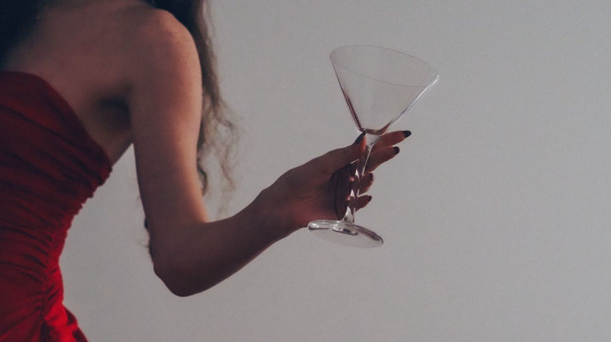 A woman is partially shown wearing a high-end red, strapless dress while holding an empty martini glass.